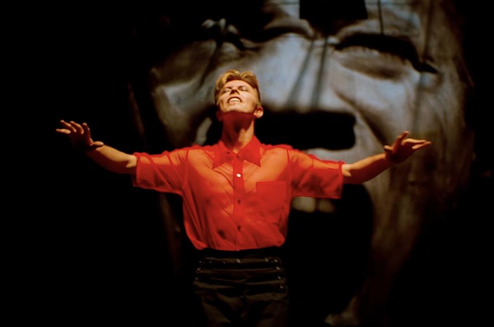 David Bowie strikes a pose in front of a black-and-white video of an agonized face in a still from MOONAGE DAYDREAM.