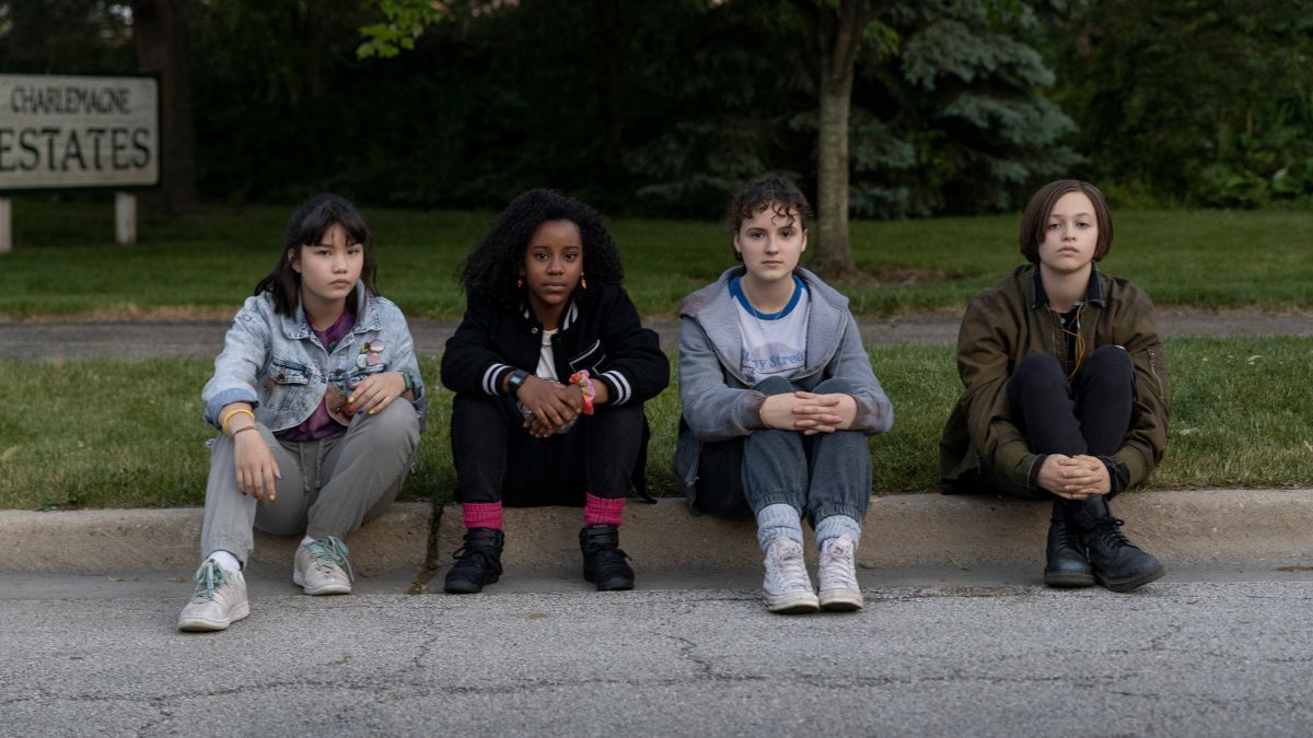 PAPER GIRLS' Riley Lai Nelet, Camryn Jones, Fina Strazza and Sofia Rosinsky sit on a curb, in character.