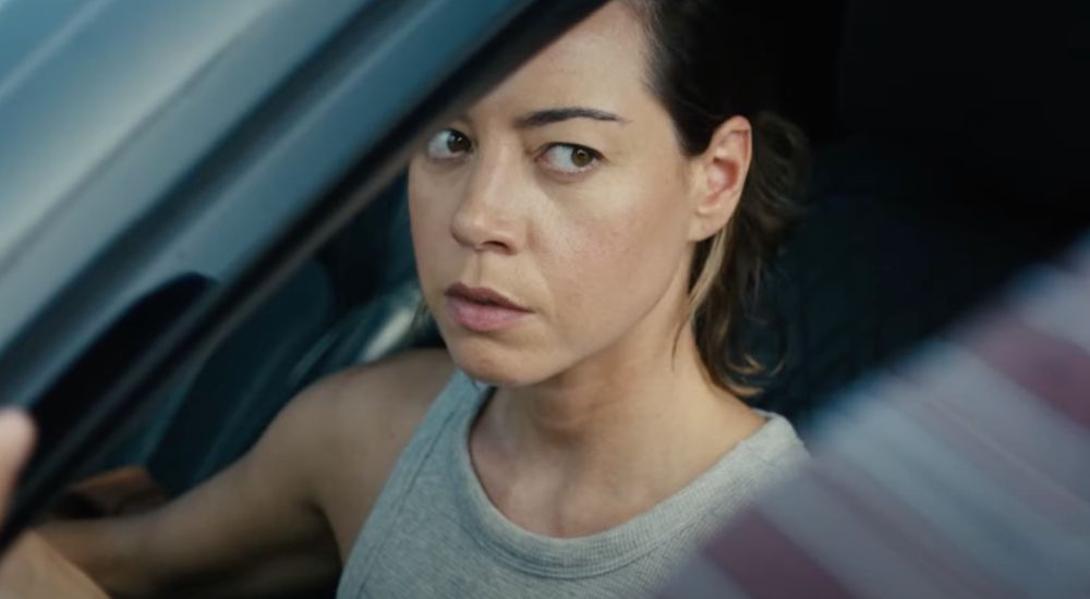 Aubrey Plaza peers suspiciously through a half-open car window in a still from EMILY THE CRIMINAL.